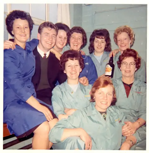 East Riding Museums A retro photograph showing eight women and a man gathered closely together and facing the camera. Most wear light blue work coats, suggesting a workplace setting, while the man near the centre wears a dark suit and tie. The group is arranged in two rows, with some seated at the front and others standing behind, resting hands on shoulders. One woman holds a glass bottle. The background includes pale tiled walls and a window frame.