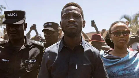 Bobi Wine in a black shirt flanked by a police officer and his wife in a blue shawl on voting day.
