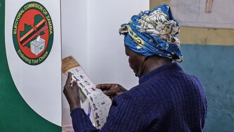 A person stands inside a polling booth in a classroom-like setting, marking a ballot. The room has worn walls, wooden desks, and hand‑drawn posters displayed on the walls. 
