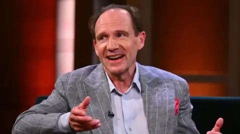 PA Media Ralph Fiennes during filming for the Claudia Winkleman Show. He is sitting on a chair and wearing a grey blazer with white pinstripes over a light blue shirt. His hands are rested in conversation. 