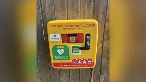 Southport Saviours Foundation A defibrillator - a medical device which is yellow, green and red - mounted on a wooden panel. 