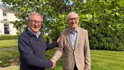 Guernsey Community Savings founder and now life president Peter Neville (left) with new chair James Ellis. Neville is wearing a navy fleece and glasses. Ellis is wearing a light brown blazer and blue shirt and glasses. The pair are shaking hands, looking at the camera and smiling. 