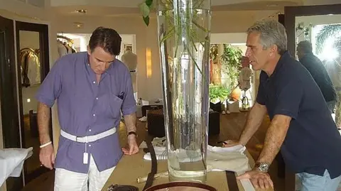 United States District Court Southern District of New York Peter Mandelson and Jeffrey Epstein pictured together. Mandelson, on the left of the photo, wears a purple short-sleeved shirt, a white belt and white trousers. He has short brown hair and is looking down. Epstein, on the right, is looking at him from across a small table, on which he is leaning and from which a large glass tube stretches upward. He is wearing a navy polo shirt, blue jeans and a large watch. He has short grey hair. The kind of room they are in is unclear - the walls are white, there are dark brown doorways and there is various clutter in the background, with some leafy plants visible.