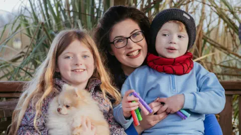 The Kennel Club/Elle Riley A family, including Rozalyn, mum Abbie and Rory, are sat outside smiling. Rosa the Pomeranian is being held by Rozalyn on the left hand side. 
