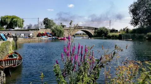 Rubytwoshoes The River Thames in Lechlade and the halfpenny bridge above it. Image focuses on pretty purple flowers with water and bridge in the background.