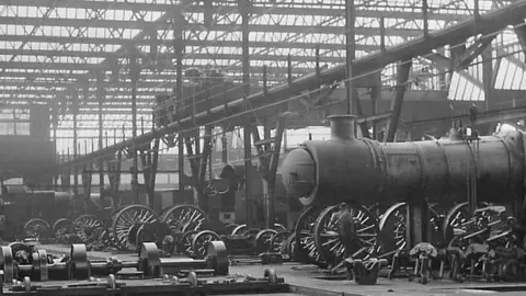 Swindon Borough Council An old black and white photo taken of the railway works, showing steam engines lined up and under construction in a large warehouse. 