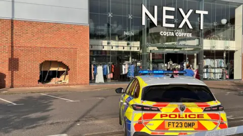 Eric Johnson A police car is parked inside a cordon in front of a large Next store. The the left of the shop window is a large hole in a brick wall where a car had crashed through.