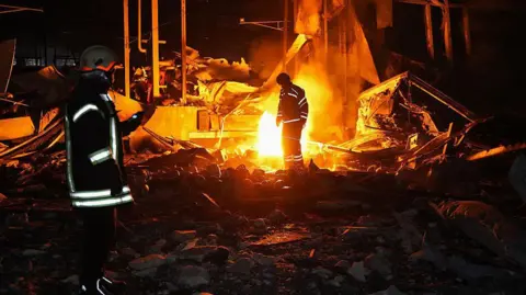 Emergency workers stand in front of a blaze at night. reflective strips on their coats stand out in the shadows and light from the fire.