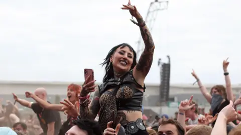 A woman stood on the shoulders of a man with her left hand in the air with other people behind her also standing on people's shoulders enjoying music at the Reading Festival 2025.