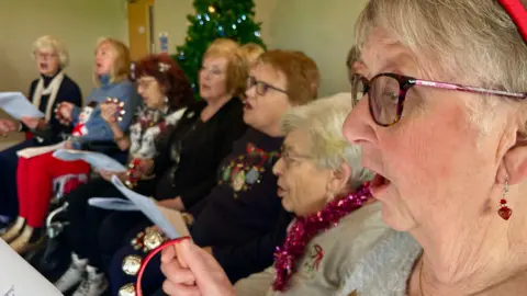 Martin Giles/BBC Seven women sitting in a line, singing. They all holding music sheets, with some of them looking down at the music and some looking straight in front of them.