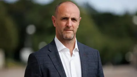 PA Media A man stares into the camera. He has balding dark hair and stubble on his face. He is wearing a dark suit jacket and open collared white shirt. The background is green and blurred.
