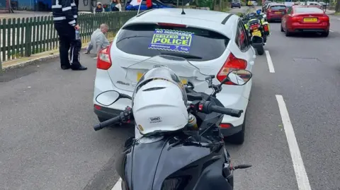 A sign reading "No Insurance, seized by police" at the back of a car. A police offer is seen nearby with a citizen who sits on the pavement. Other people are near them. Cars keep driving on the road.