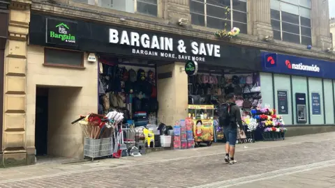 A shop called 'Bargain and Save' which is in an historic building but has lots of items outside like brooms and plastic boxes and flowers. There is a nationwide bank next door. 