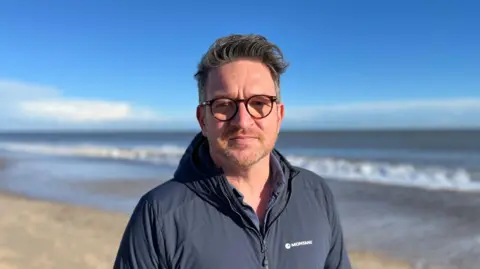 Shaun Whitmore/BBC Prof Ben Garrod, wearing glasses and a navy jacket, standing on the beach. The sea can be seen behind him.