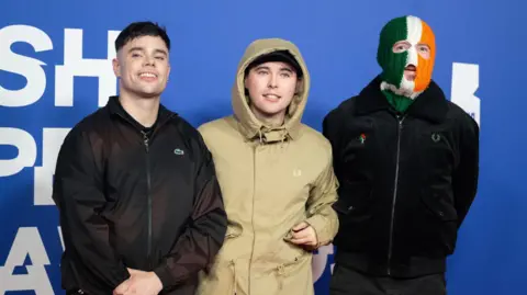 Getty Images Móglaí Bap, Mo Chara and DJ Próvaí walk the red carpet at the British Independent Film Awards. DJ Próvaí on the right is wearing an Irish tricolour balaclava.