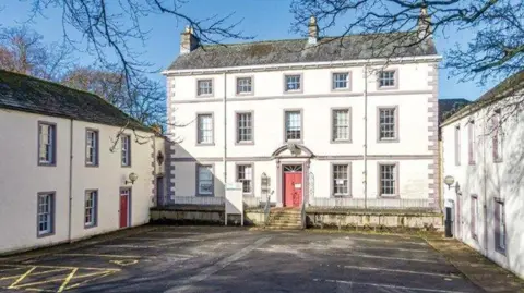 Mansion House consists of three white buildings in a u-shape. The main building is three-storey tall with rows of square windows and a staircase leading up to a large red door. The buildings on the left and right are two-storeys with a similar colour design. There are car parking spaces marked in paint out front.