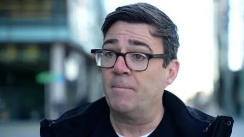 Andy Burnham speaking to camera. He had short black hair, black glasses, and is wearing a dark jacket