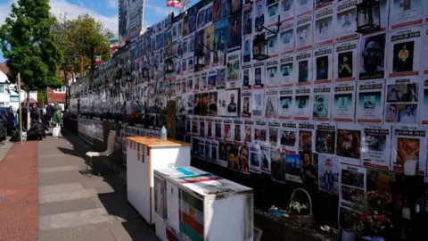 A wall of people's faces and information is topped by Iranian flags. Flowers and furniture also line the wall.