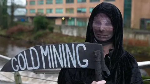 A protester is dressed up like Death. They are wearing a black hooded cape and is holding a scythe that says goldmining. Their face is obscured. A river is behind them. 