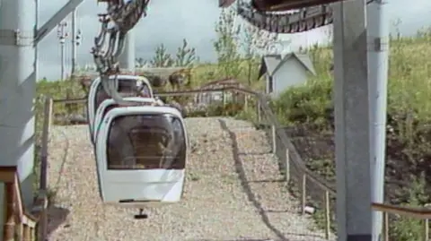 Three cable cars can be seen, front-on, surrounded by ornamental gardens. The cable car gondolas are white, with a large pillar sticking up out of the ground to hold the cables and wheels of the system.