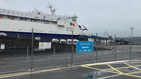 Shows a large ferry with a lighthouse in the background and a blue and white signpost saying Cairnryan