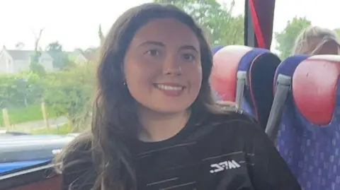 Handout Ellie McBride with dark hair, she is smiling at the camera. She is wearing a black sports top. Looks like she is on a bus or coach.