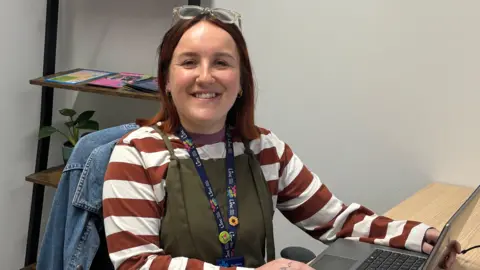 Ellie smiles at the camera while working at a laptop in an office. She is wearing green dungarees, a brown and white striped top and has clear-framed glasses perched on her head. 