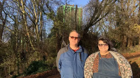 A man with short cropped hair is wearing glasses and a blue and grey anorak. He is stood with a woman with long black hair with a green streak. She is wearing a patterned cardigan, a leopard print top and dungaree dress. They are stood in front of a tower block and woodland