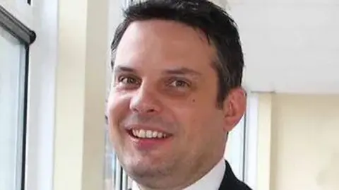 Christos is smiling at the camera against a cream wall. He is wearing a white shirt and navy suit jacket. He has brown hair and hazel looking eyes. 