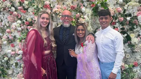 Family handout A group of four people stand together in front of a wall covered in flowers.
