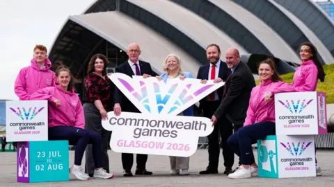 PA Media Athletes in pink hoodies flank official figures who are standing outside the SEC Armadillo in Glasgow holding Commonwealth Games branding. They are, from left, Glasgow City Council leader Susan Aitken; First Minister John Swinney; Commonwealth Games Federation chief executive Katie Sadleir; Scottish Secretary Ian Murray and the chief executive of Commonwealth Games Scotland Jon Doig.