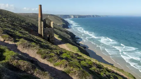 Adam Gibbard/Visit Cornwall A derelict engine house sits on a steep cliff above a long pale sandy beach and the rest of the coast stretches out into the distance, alongside high rugged cliffs. The sea and sky are blue and it is sunny.