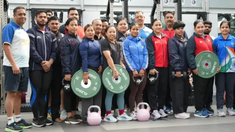 The men and women of the Indian weightlifting team lined up in a gym to pose for a photo, They are wearing gym clothes and some are holding large, round weights.