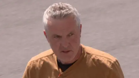 Brendan O'Meara. He has short white hair, wearing a mustard coloured top.