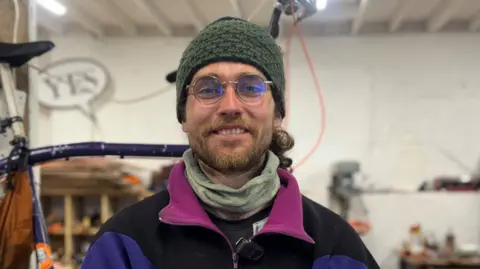 Alan is smiling at the camera. He is standing in a workshop with his tall bike behind him. He is wearing round glasses, a green beanie hat and a colourful jumper.
