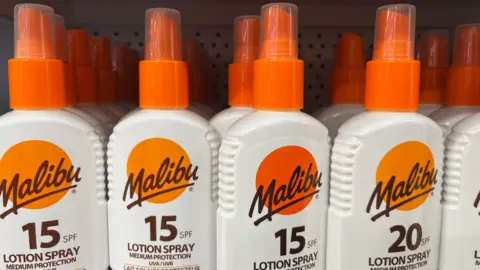 A row of Malibu SPF 15 sun-tan lotion sprays on a shelf. They have an orange lid and a white body, with branding.