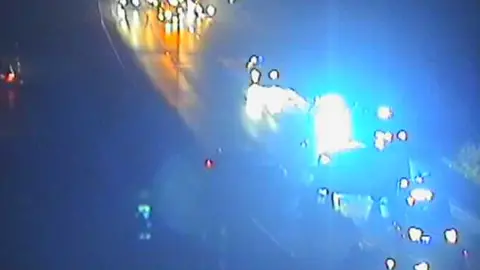 Highways England A still from a CCTV camera overlooking the motorway. It is blurry, but the blue lights of emergency vehicles are in the image and have blocked off the road. A queue of traffic waits behind them.