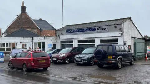 Forecourt of Urchfont Vehicles garage with four cars near a road.