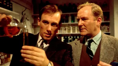 Two men look at a glass vial as one pours it in a picture from the 1980s