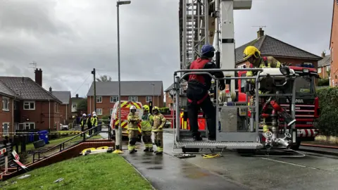 Two fire officers are in an aerial ladder which is attached to a fire engine. There are fire engines standing on a pavement nearby. There is tape cordoning off a building