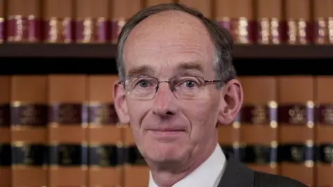 Royal Courts of Justice Sir Andrew McFarlane berdiri di depan baris buku coklat. Dia memiliki rambut pendek beruban, kacamata tanpa bingkai dan dicukur bersih