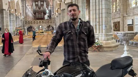 Abraham Saxon standing behind his black motorcycle inside the cathedral. He has short dark hair and and is wearing a dark coloured shirt. 