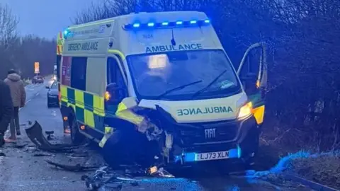 People stand in the middle of the road including one man on a phone at the scene of a car crash. A grey car with a logo is across one lane and partly on the verge. The ambulance, which has been damaged, is on the other side of the road, with one door open. Other cars are around the crash scene and it is twilight. There are white lines in the middle of the road.