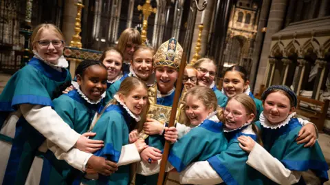 Finnbarr Webster A dozen girls in the cathedral wearing chorister uniform of white undercoats with blue tunics on top, all smiling to camera and hugging Emmie Piper who is wearing vestments similar to a bishop and a mitre