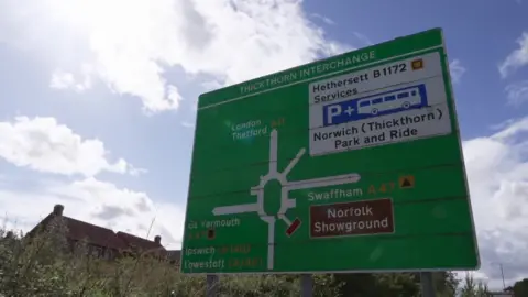Emma Craig/BBC Large road sign at Thickthorn Roundabout on the A11. Roofs of houses can be seen in background.