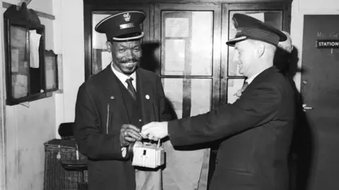 Getty Images A black man, smiling and wearing a smart uniform is handed a container by a white man in similar uniform.