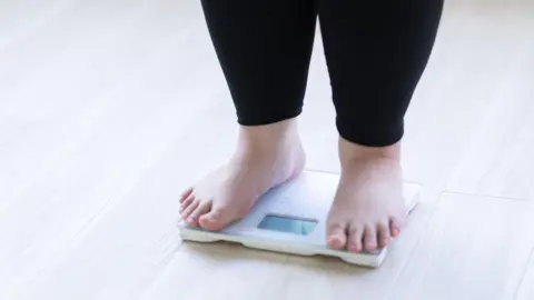 A child stands on weighing scales wearing black leggings and bare feet