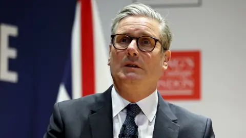 Keir Starmer looks on as he speaks at an event in London on Thursday. He's wearing a suit and tie with glasses on.