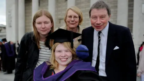 Lucinda Ritchie Lucinda sits in a wheelchair with her graduation cap and gown, surrounded by three members of her family. 