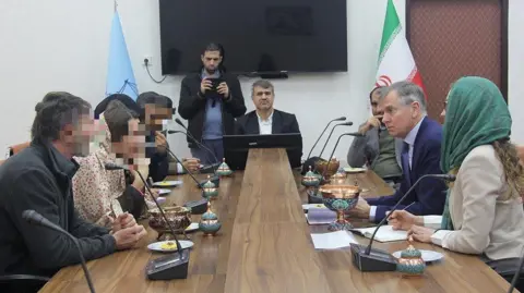 IRNA Several people sit around a wood-panelled table - laid with with ornate bowls and pots. Hugo Shorter, a man wearing a blue suit and tie, sits on the right hand side between a woman wearing a green scarf over her hair and a man in a grey jacket and green shirt. On the left hand side are four people with their faces blurred while at the far end there are two men wearing black coats, one of who is holding a mobile phone